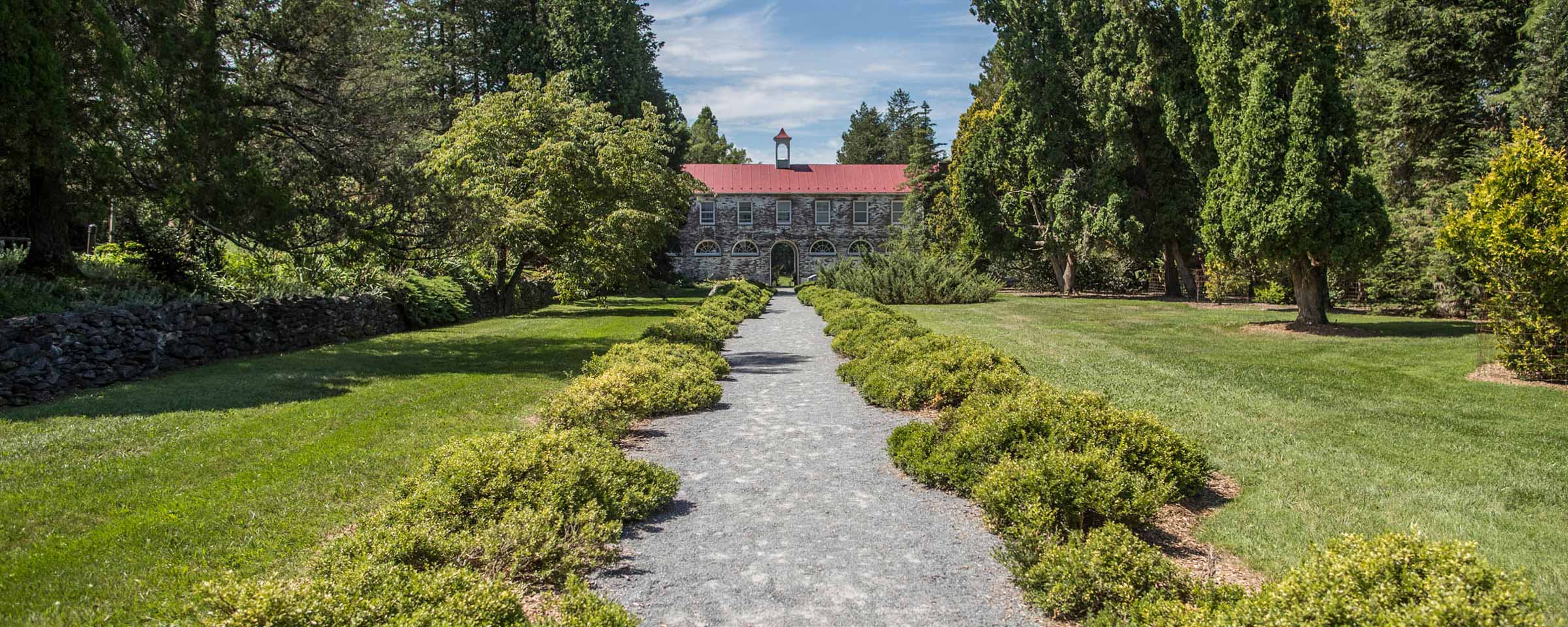 Two story stone building with a red roof at the end of gravel sidewalk lined with shrubs